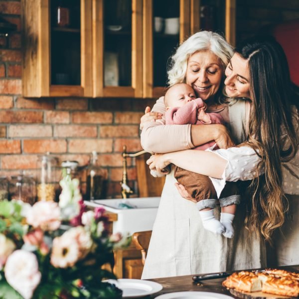 Joyful aged lady plays and hugs with little granddaughter and daughter near brown wooden kitchen.